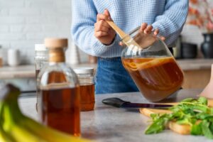 A person in a blue sweater stirs a jar of brown liquid, surrounded by bottles