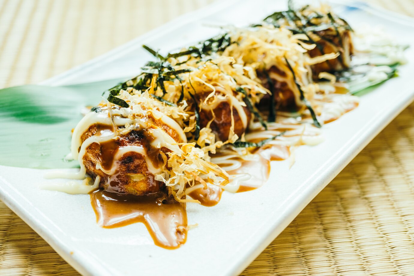 Plate of Takoyaki with toppings, drizzled with sauce on a white rectangular dish, served on a woven mat.
