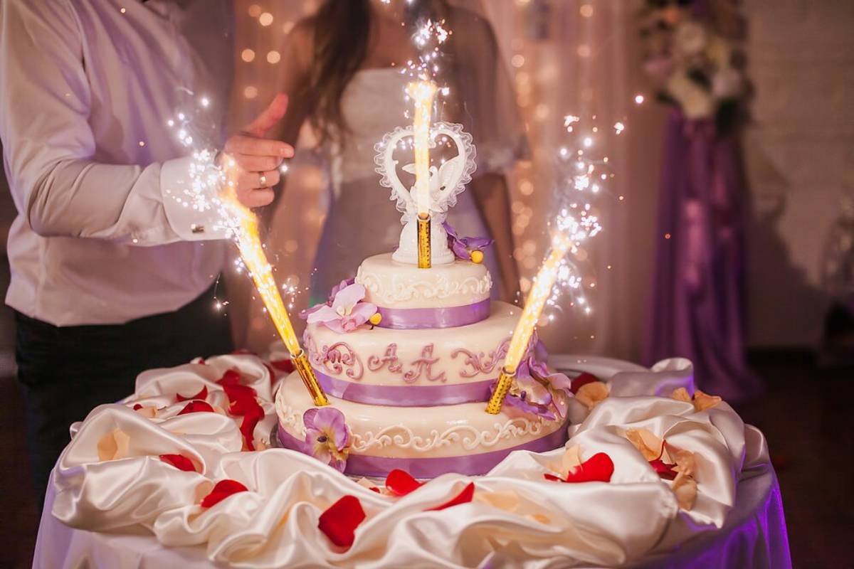 A couple holding sparklers by a decorated wedding cake with a heart-shaped topper.