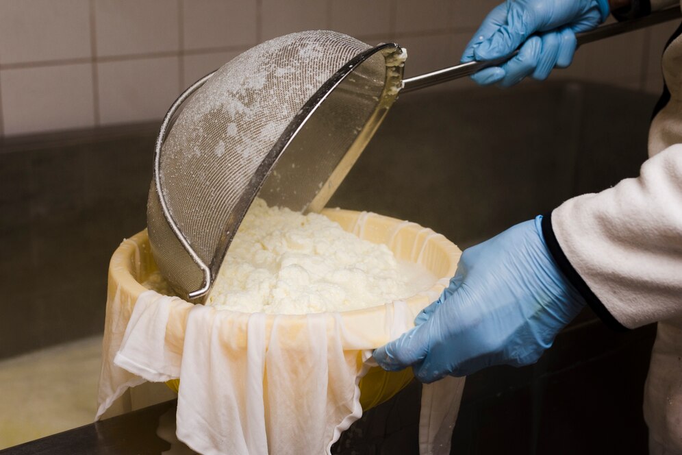 Person in gloves drains cheese curds through a mesh sieve into a cloth-lined bowl.