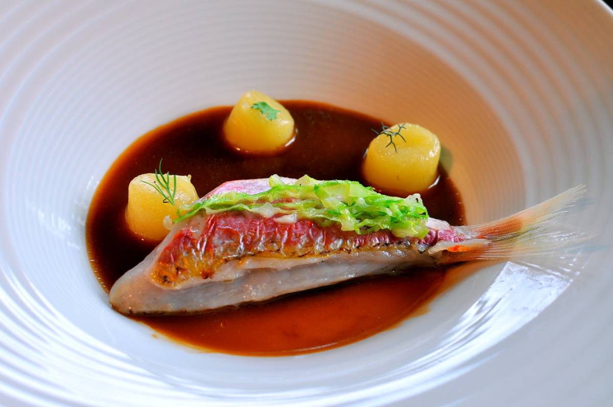 A gourmet dish featuring a whole fish with crispy skin, served with round yellow potatoes and a rich, dark sauce in a white bowl.