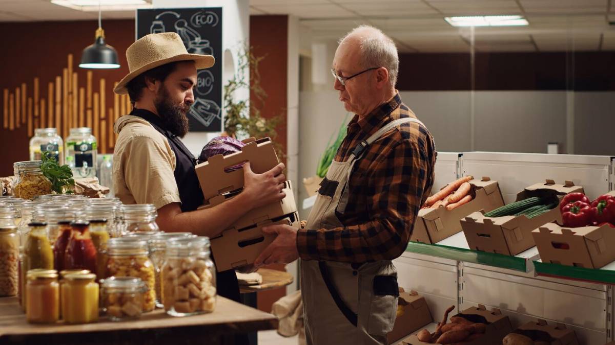 Two men exchanging produce boxes in a small, cozy grocery store with shelves stocked with various preserved foods.