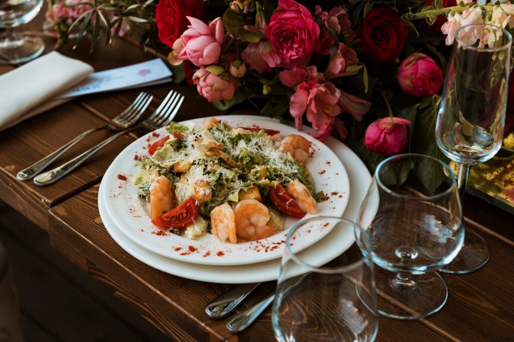 A gourmet shrimp salad served on a white plate beside a floral centerpiece and wine glasses on a wooden table.