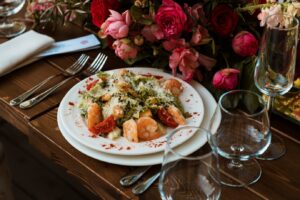 A gourmet shrimp salad served on a white plate beside a floral centerpiece and wine glasses on a wooden table.