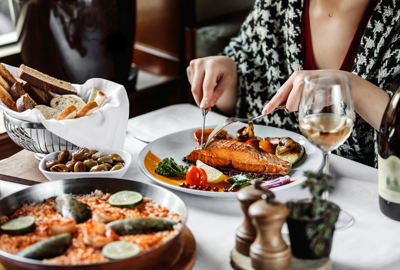 Woman in a restaurant cuts into a salmon filet on her plate, surrounded by various dishes and a glass of white wine.