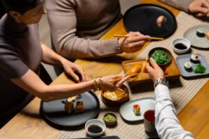 Three people dining at a sushi restaurant, sharing sushi and side dishes on a wooden table.