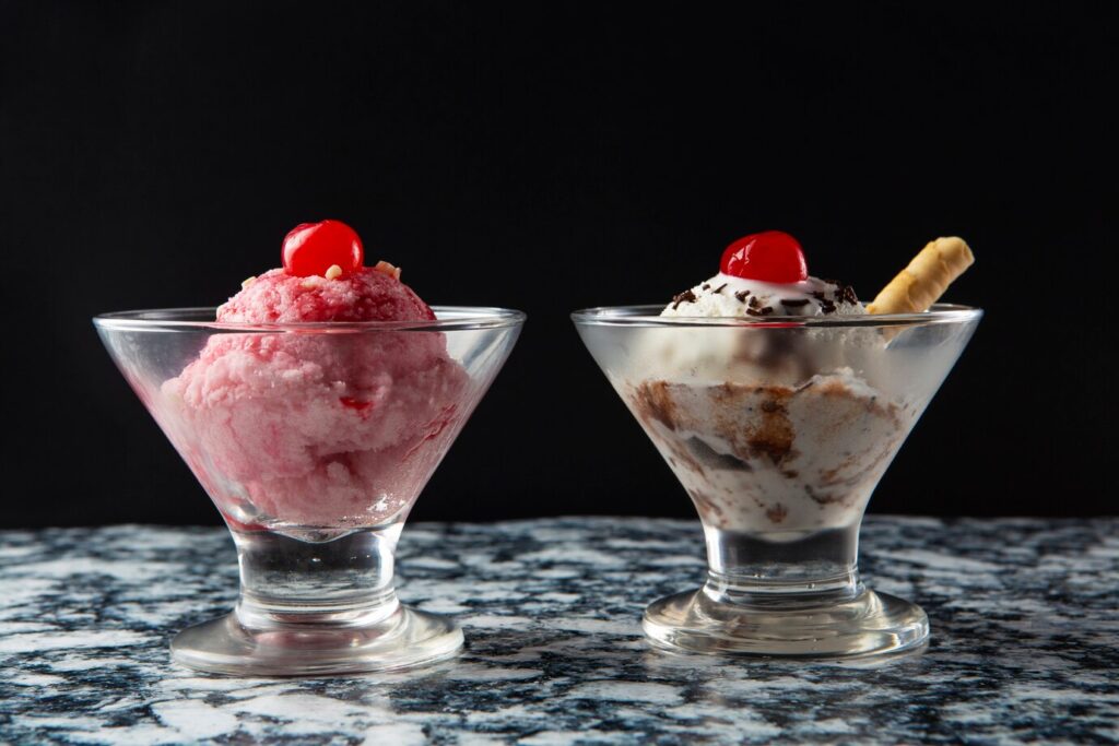 Two bowls of ice cream on a marble surface; one pink with a cherry, one white with a cherry and wafer.
