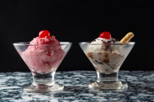 Two bowls of ice cream on a marble surface; one pink with a cherry, one white with a cherry and wafer.