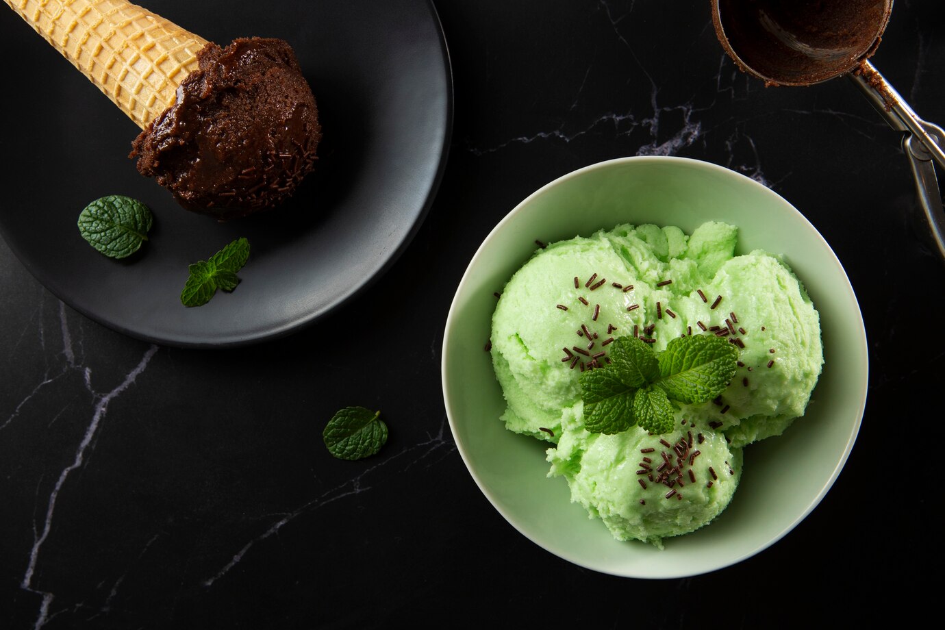 Bowls of chocolate and mint ice cream with sprinkles and mint leaves on a dark marbled surface.