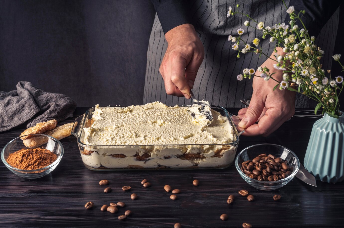 Person preparing tiramisu, spreading cream over layers in a glass dish, with ingredients and flowers nearby.