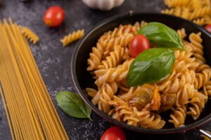 A bowl of fusilli pasta with tomato sauce, fresh basil, and cherry tomatoes on a dark surface.