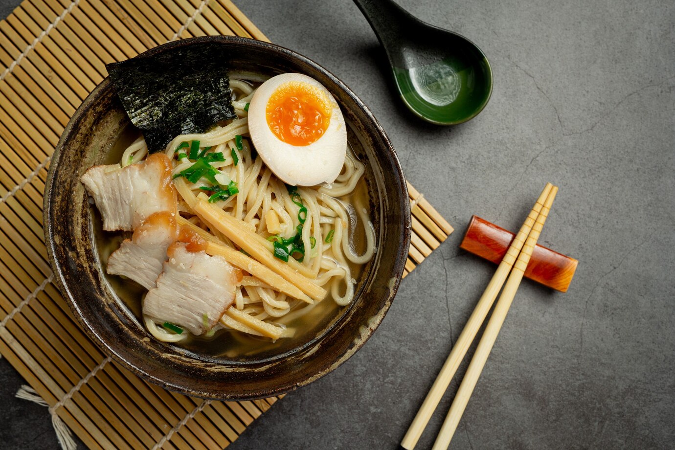 Bowl of ramen with pork, egg, bamboo shoots, and seaweed, served with chopsticks and a spoon on a gray surface.
