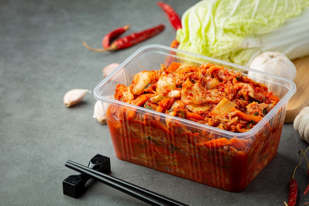 Kimchi in a clear plastic container with fresh vegetables and red chili peppers on a dark surface, black chopsticks aside.