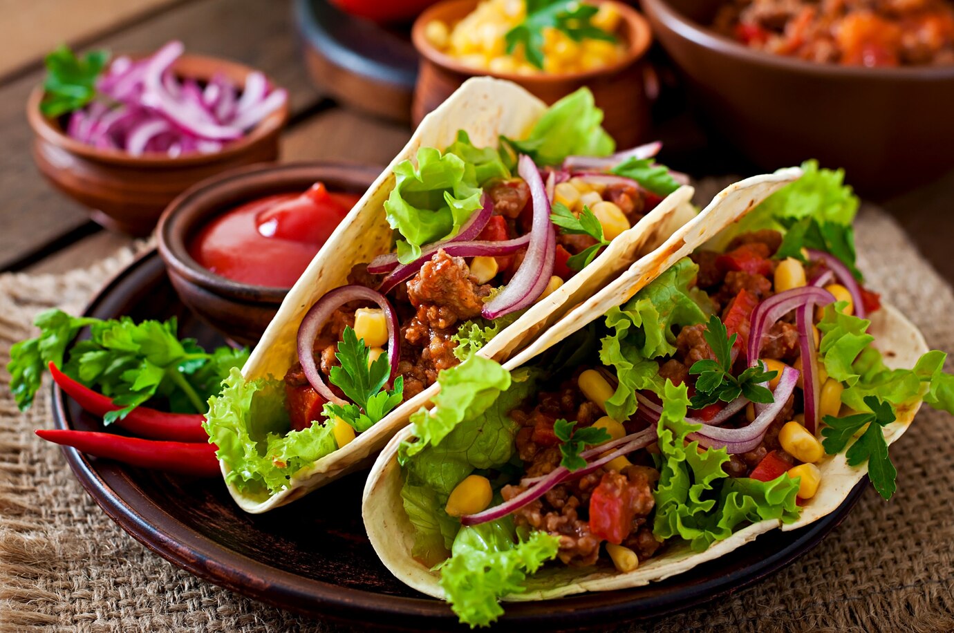 Three vibrant tacos filled with lettuce, corn, ground meat, and onions, served with condiments on a wooden table.