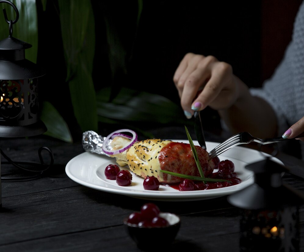 Person eating a gourmet meal with grilled chicken, cherries, and onion on a dark, elegant table setting.