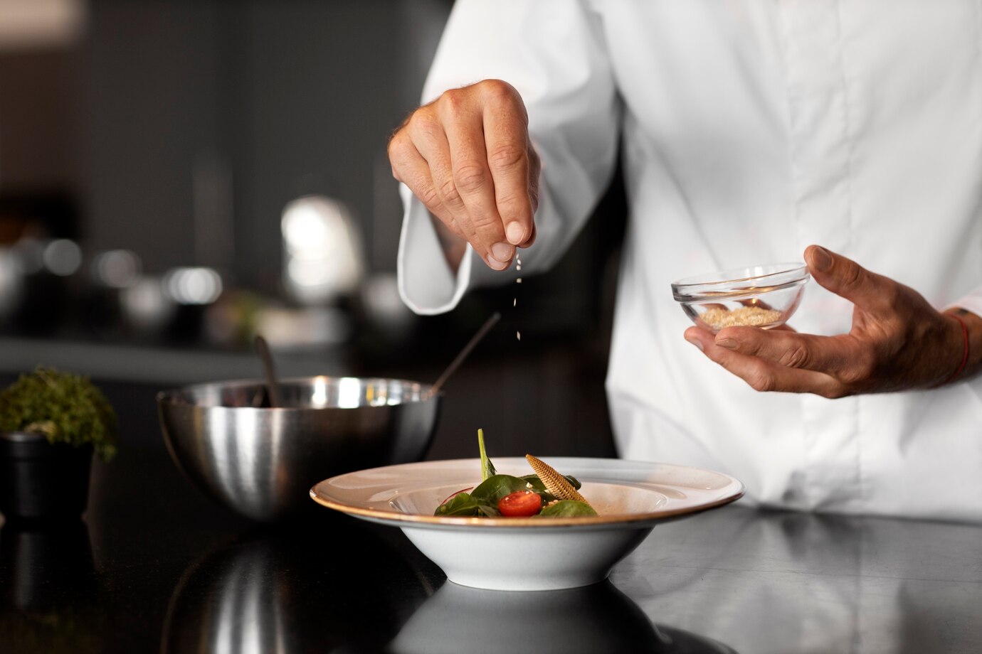 Chef in white uniform seasoning a dish with sesame seeds in a modern kitchen.