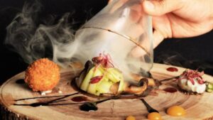 A hand lifting a smoke-filled glass dome from a creatively presented dish on a wooden platter.