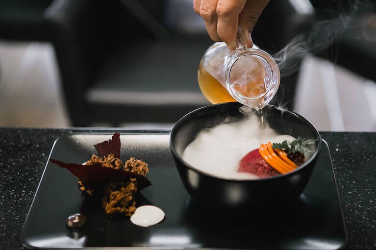 Hand pouring a clear liquid into a steaming bowl of gourmet food on a modern black tray.