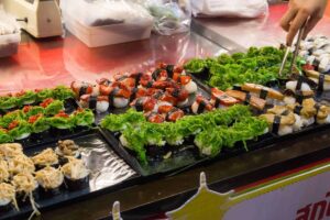 Person serving various types of sushi decorated with green lettuce at a food market stall.