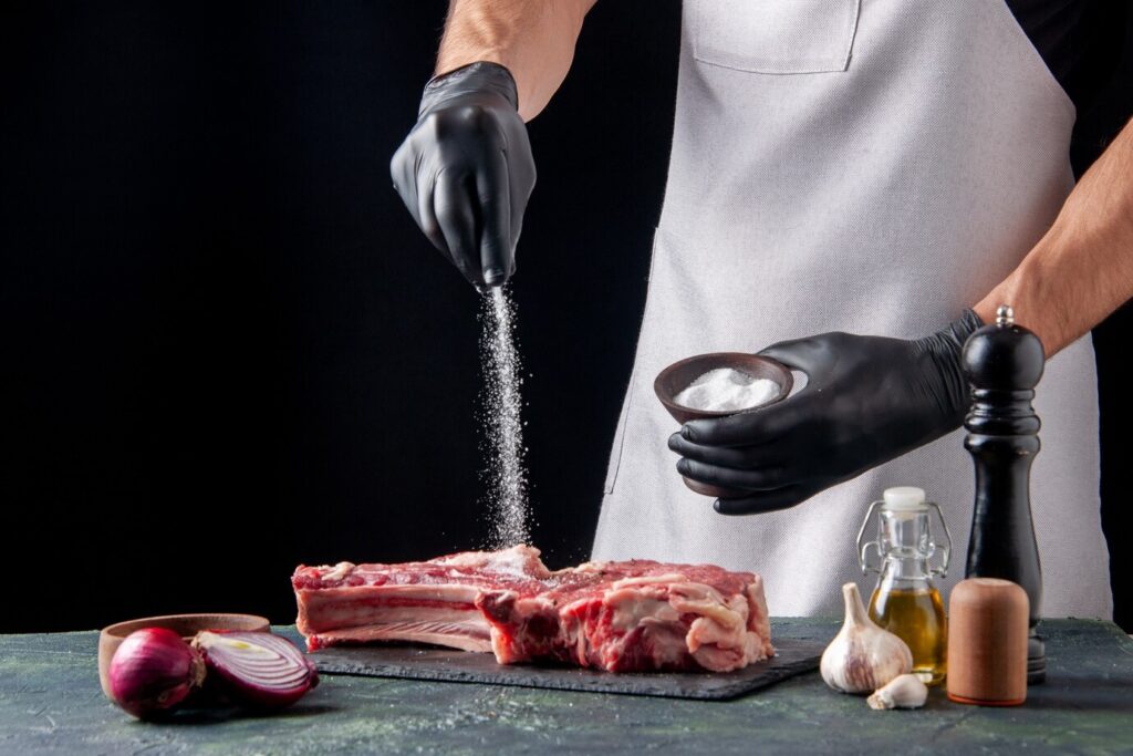 Person in black gloves seasoning raw ribs with salt, surrounded by cooking ingredients on a dark background.