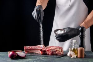 Person in black gloves seasoning raw ribs with salt, surrounded by cooking ingredients on a dark background.