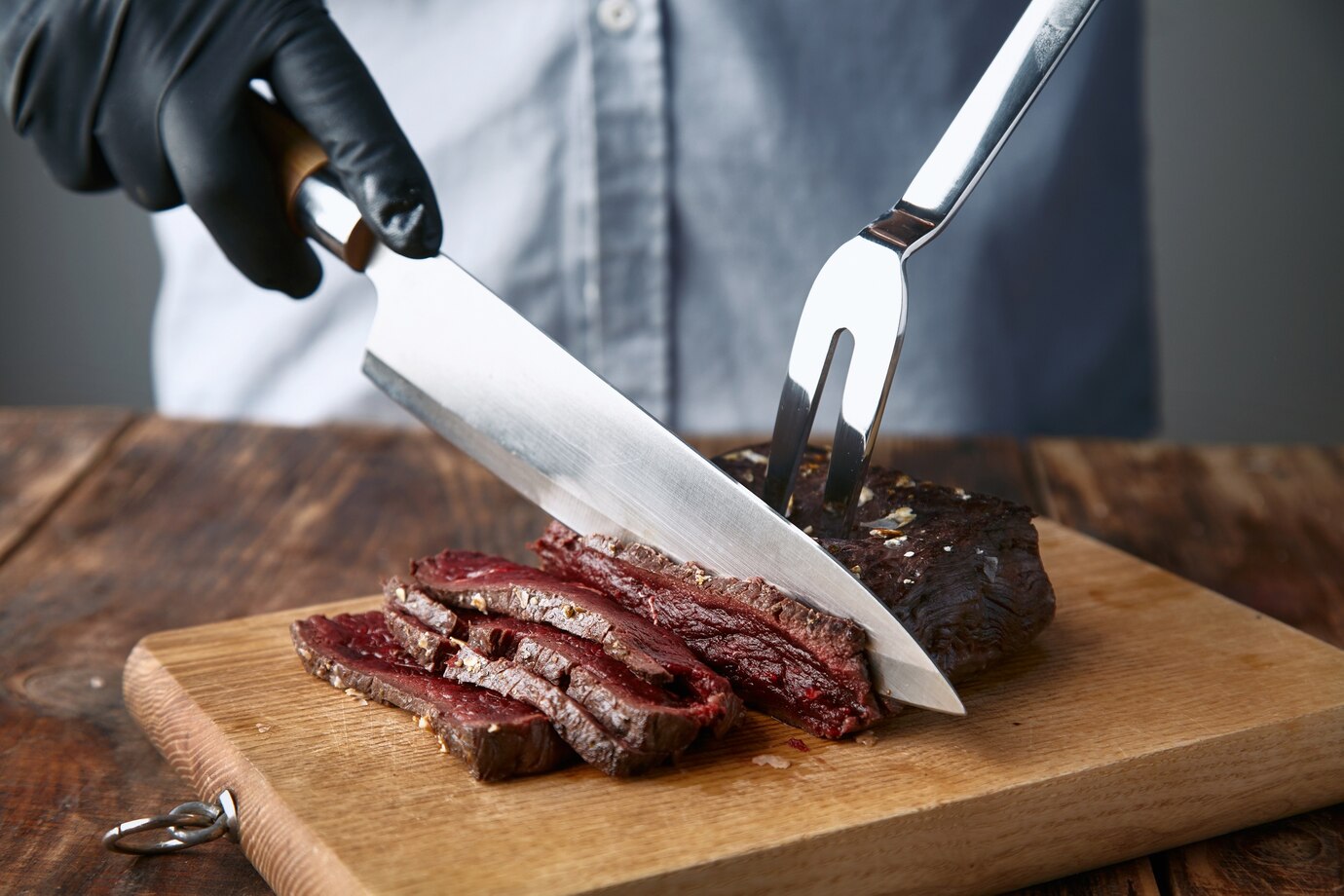 Person in black gloves cutting a cooked steak on a wooden board with a fork and knife.