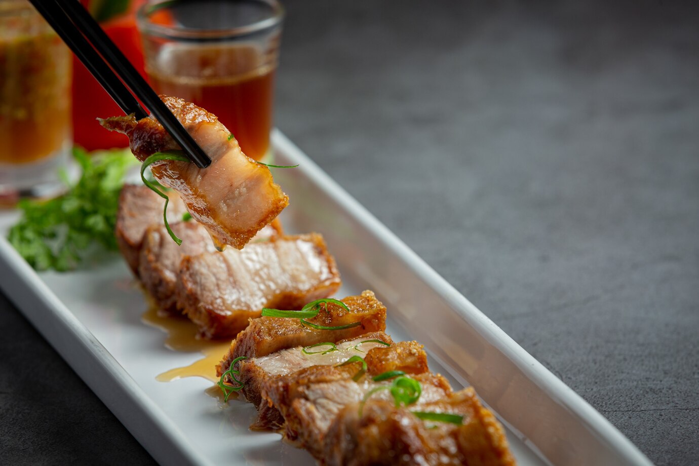 A close-up of crispy pork belly held by chopsticks, garnished with green herbs