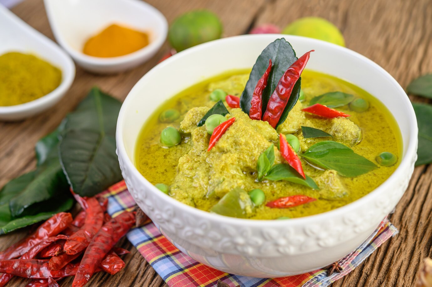A bowl of green curry with chicken, peas, red chilies, and basil leaves, served on a wooden table.