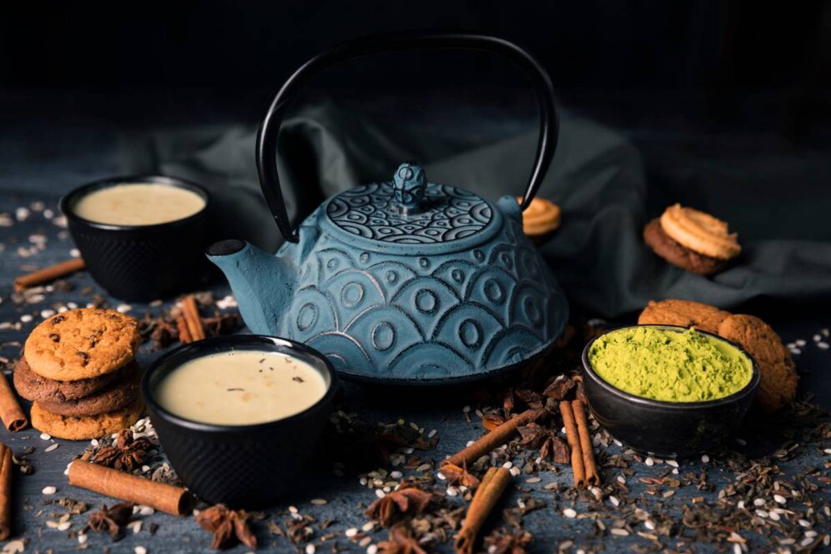 A textured blue teapot with two cups of chai, cookies, and spices on a dark background.
