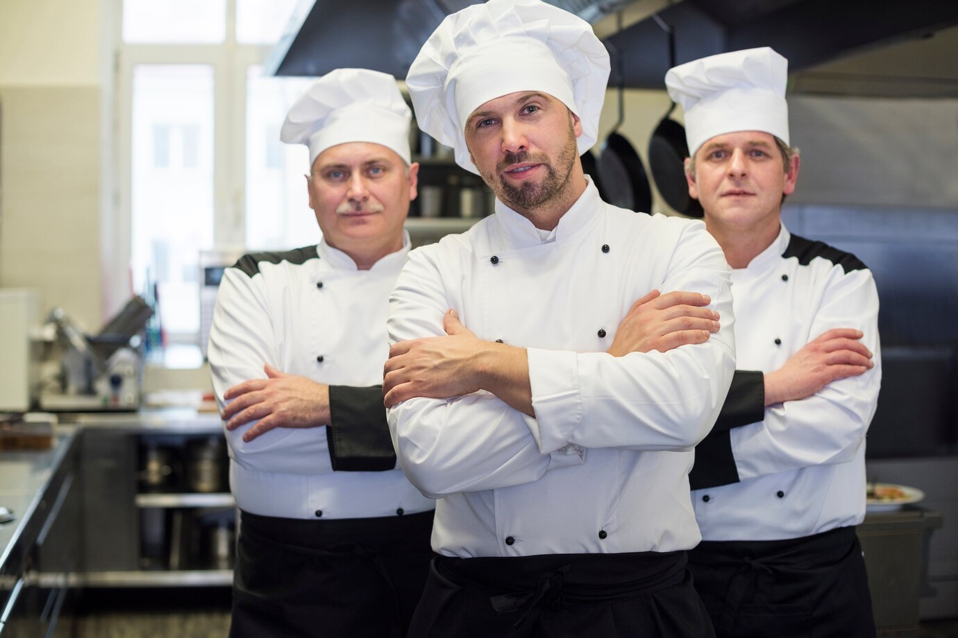 Three chefs in white uniforms and hats stand confidently in a restaurant kitchen