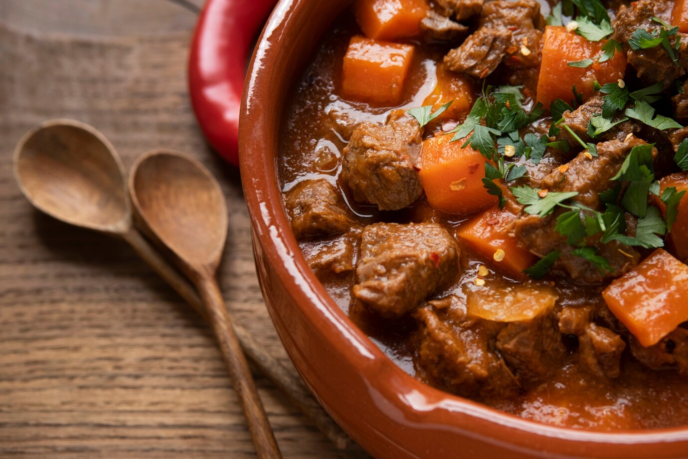 Beef stew with carrots and parsley in a clay pot, garnished with herbs, accompanied by wooden spoons.