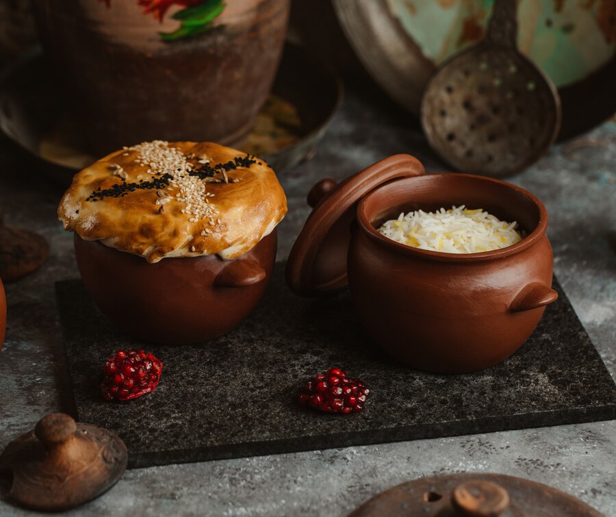 Clay pots on a table, one with a golden crusted pie, the other filled with rice, surrounded by rustic kitchenware and pomegranate seeds.