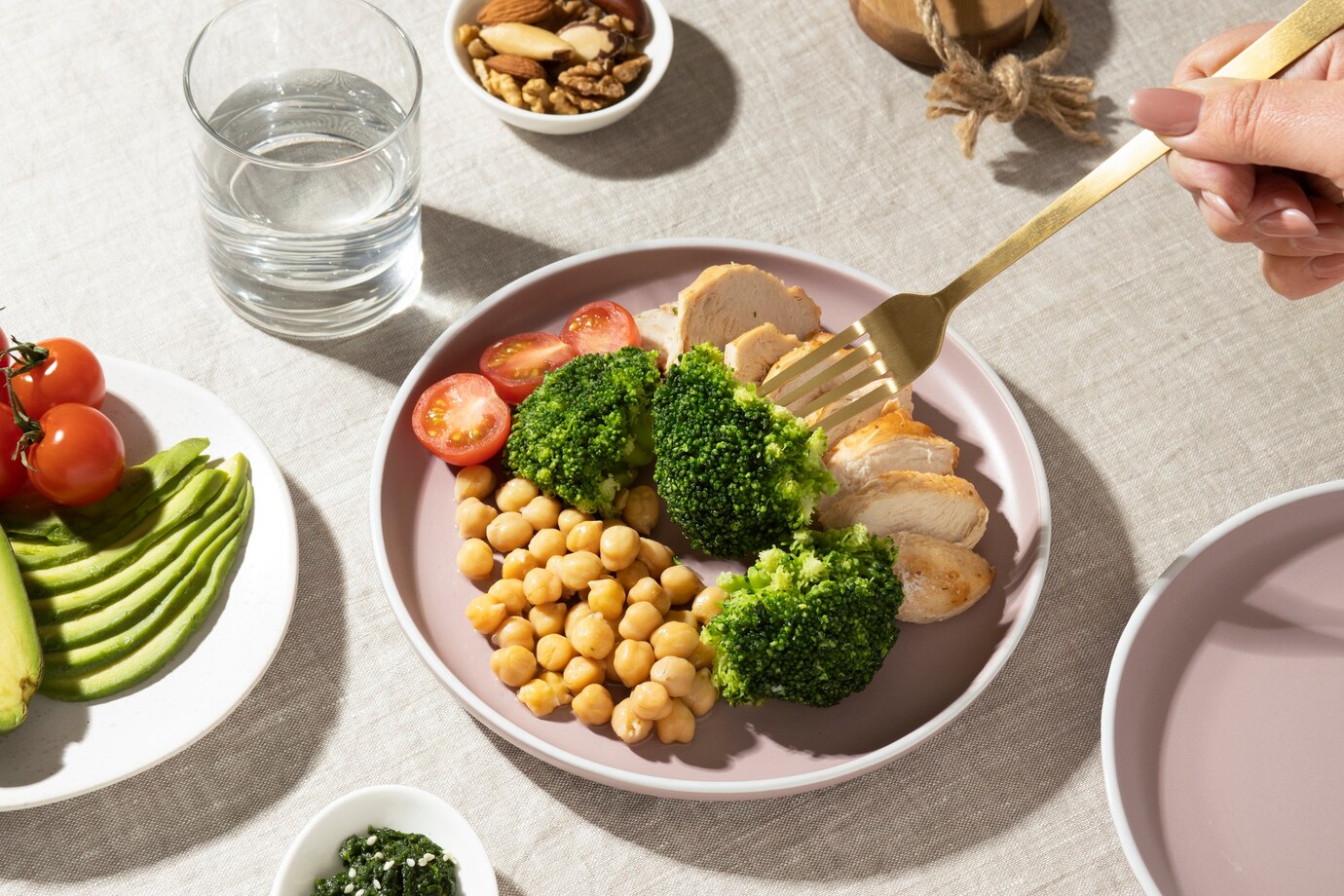 A hand with a gold fork lifts a piece of broccoli from a colorful plate featuring chicken, chickpeas