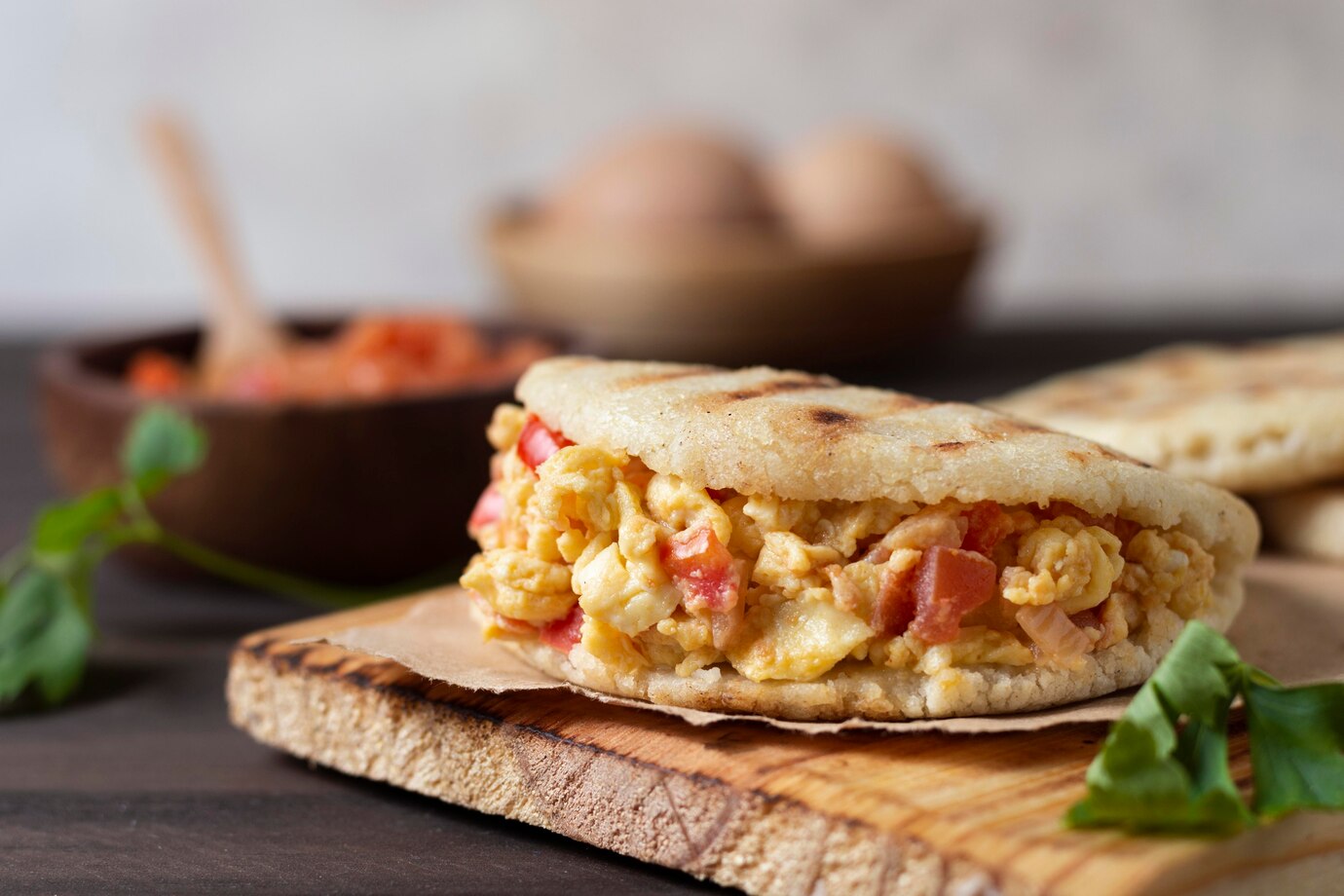 Freshly made arepa filled with scrambled eggs and tomatoes on a wooden board, with additional ingredients in the background.