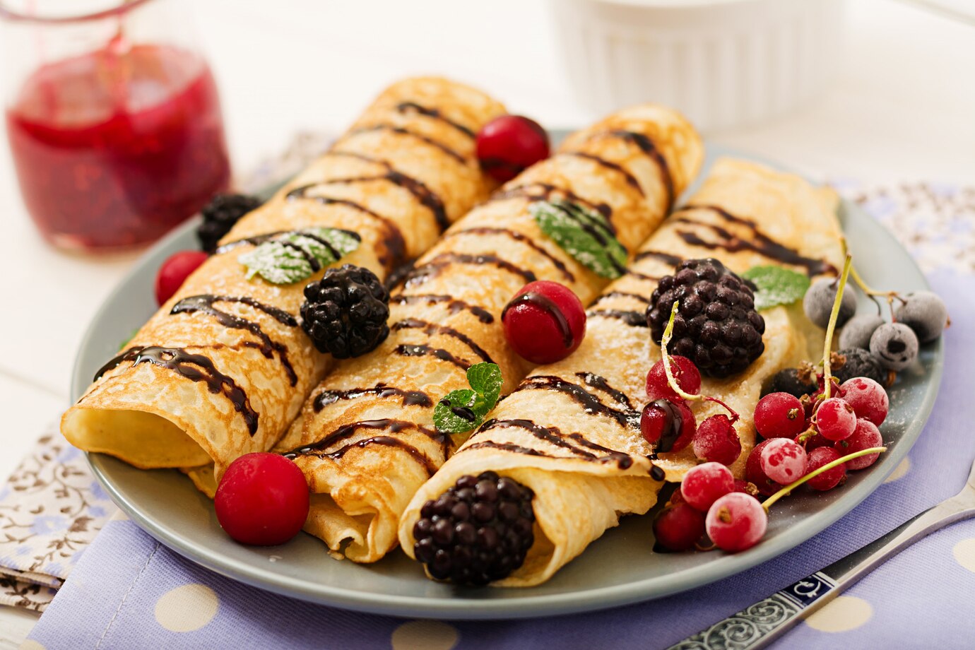 A plate of crepes topped with fresh berries, drizzled with chocolate sauce, and dusted with powdered sugar.