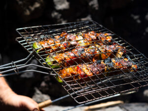 Grilled skewers with vegetables and meat on a barbecue rack held by a person's hand outdoors.