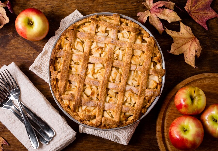 A freshly baked apple pie with lattice crust, surrounded by apples and autumn leaves on a wooden table.