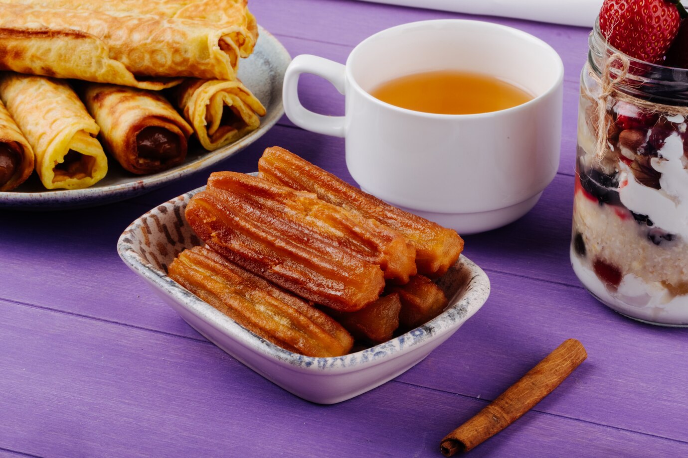 Assorted breakfast items: churros, crepes with chocolate filling, tea, and a parfait with berries and cream on a purple table.