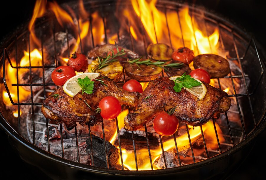 Grilled chicken legs, tomatoes, and sliced potatoes over an open flame on a barbecue.