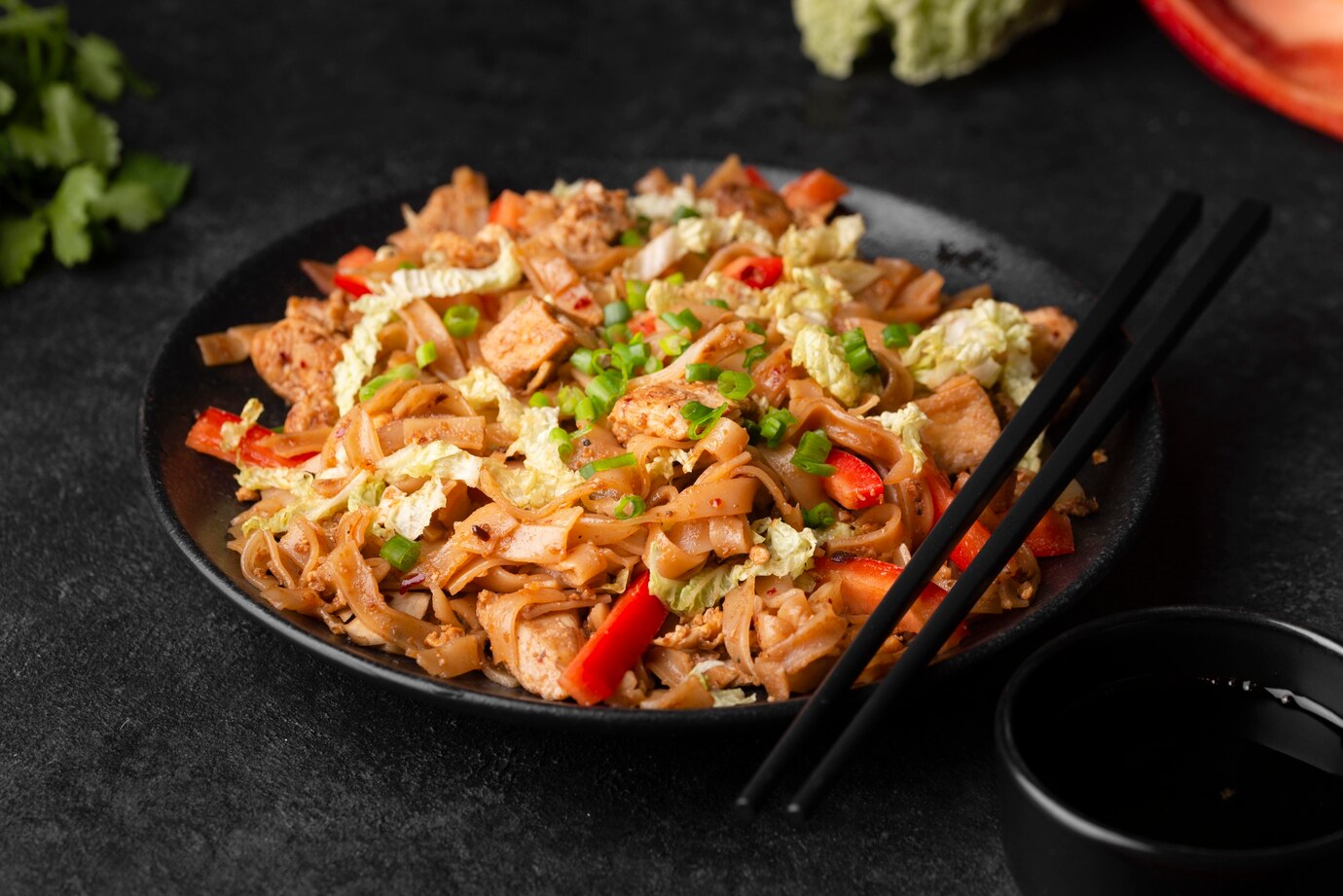 Stir-fried noodles with tofu, vegetables, and chopsticks, served on a black plate.