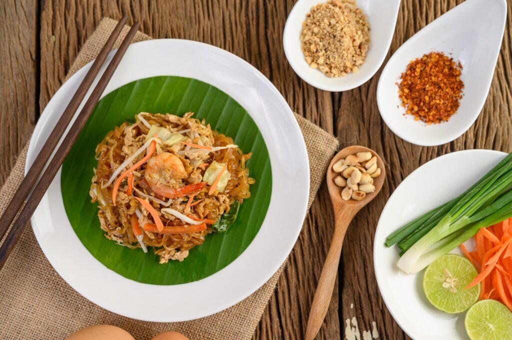 Pad Thai served on a banana leaf with shrimp, peanuts, lime, green onions, and carrots, beside various condiments.