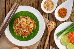 Pad Thai served on a banana leaf with shrimp, peanuts, lime, green onions, and carrots, beside various condiments.