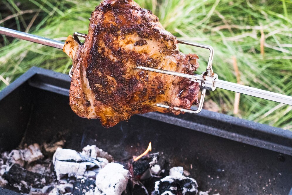 Whole chicken roasting on a spit over a charcoal grill outdoors.