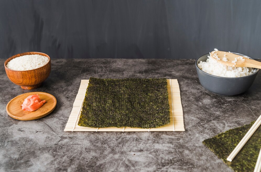 Ingredients for sushi preparation: uncooked rice in a bowl, cooked rice in another, pickled ginger on a plate, and seaweed sheets on a bamboo mat.