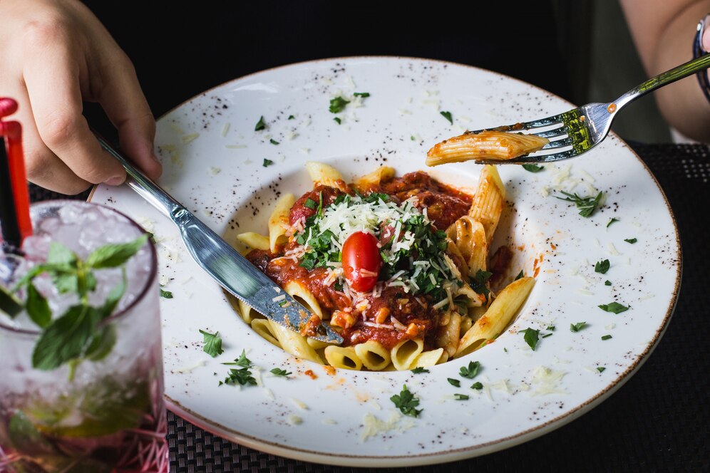 Person enjoying penne pasta with tomato sauce and cheese, served on a white plate, with a refreshing drink beside it.