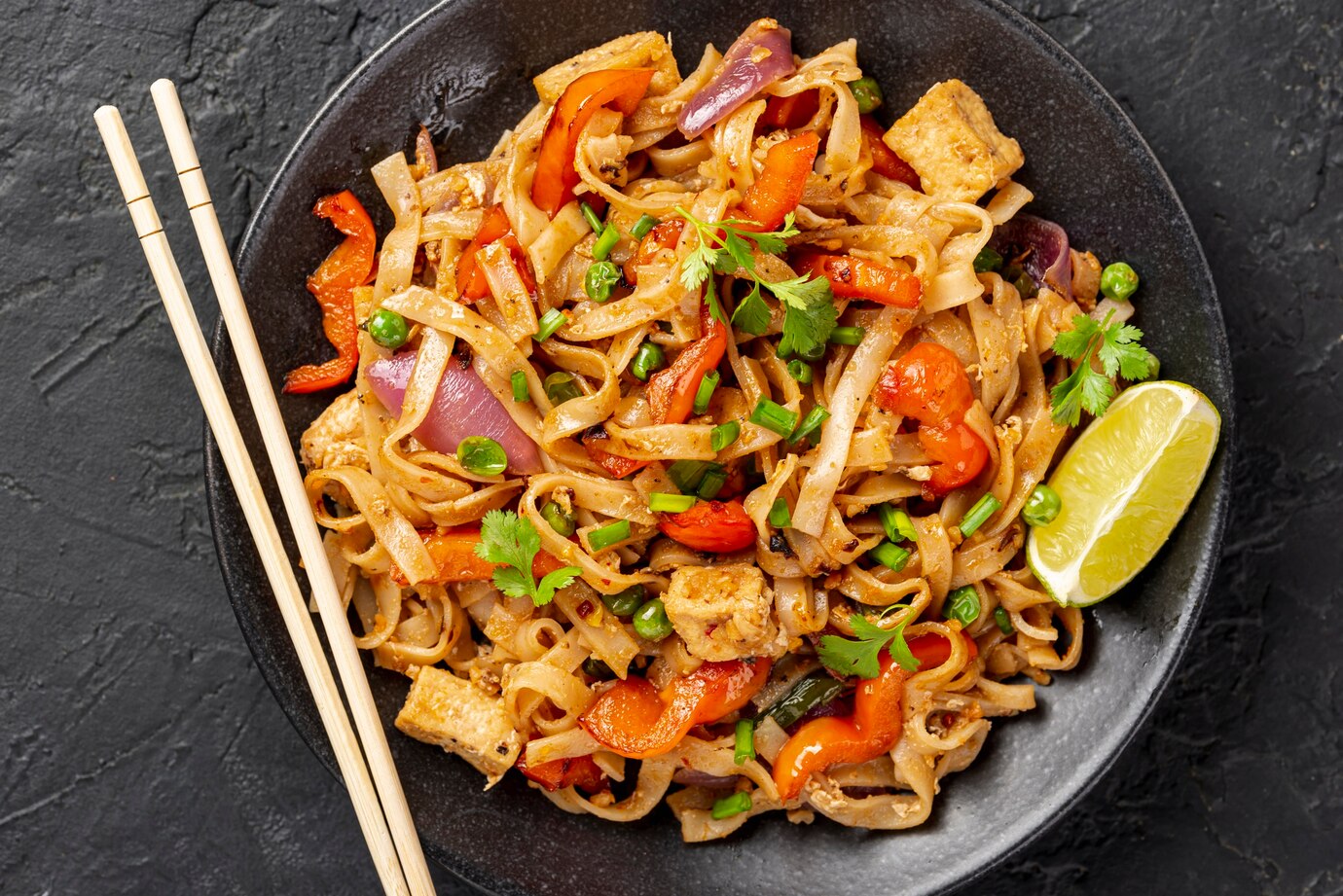 Pad Thai with tofu, vegetables, and lime in a black bowl with chopsticks.