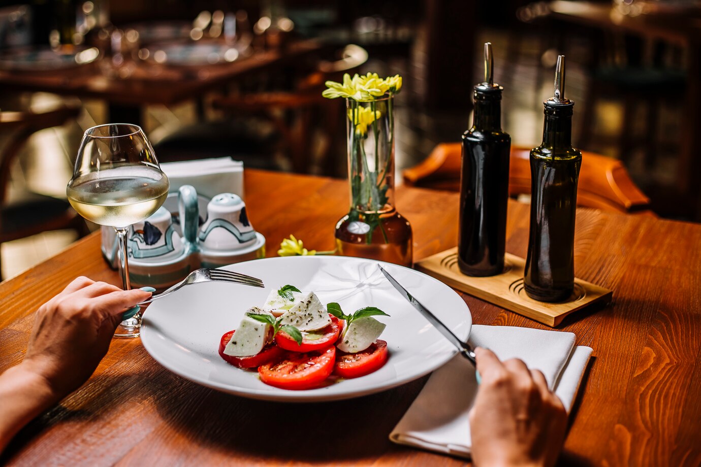 A person enjoying a tomato and mozzarella salad at a restaurant table with a glass of white wine.