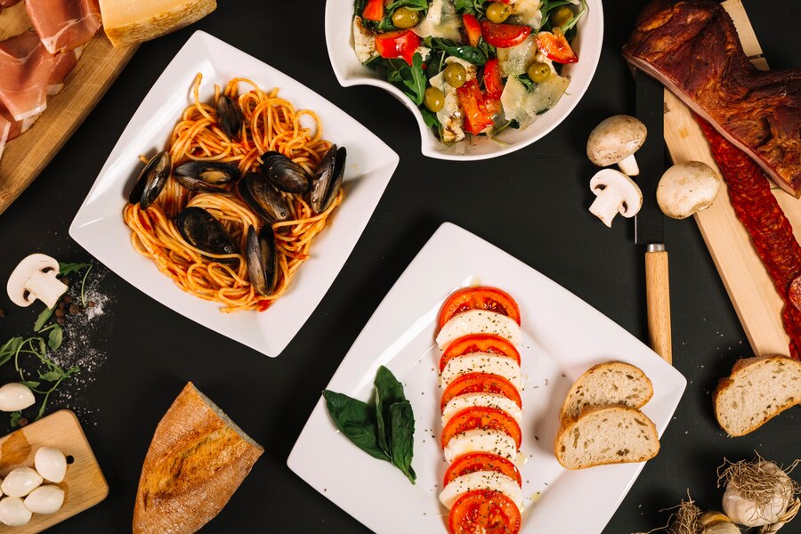A spread of Italian dishes including seafood pasta, caprese salad, and a mixed vegetable salad, arranged on a dark table.