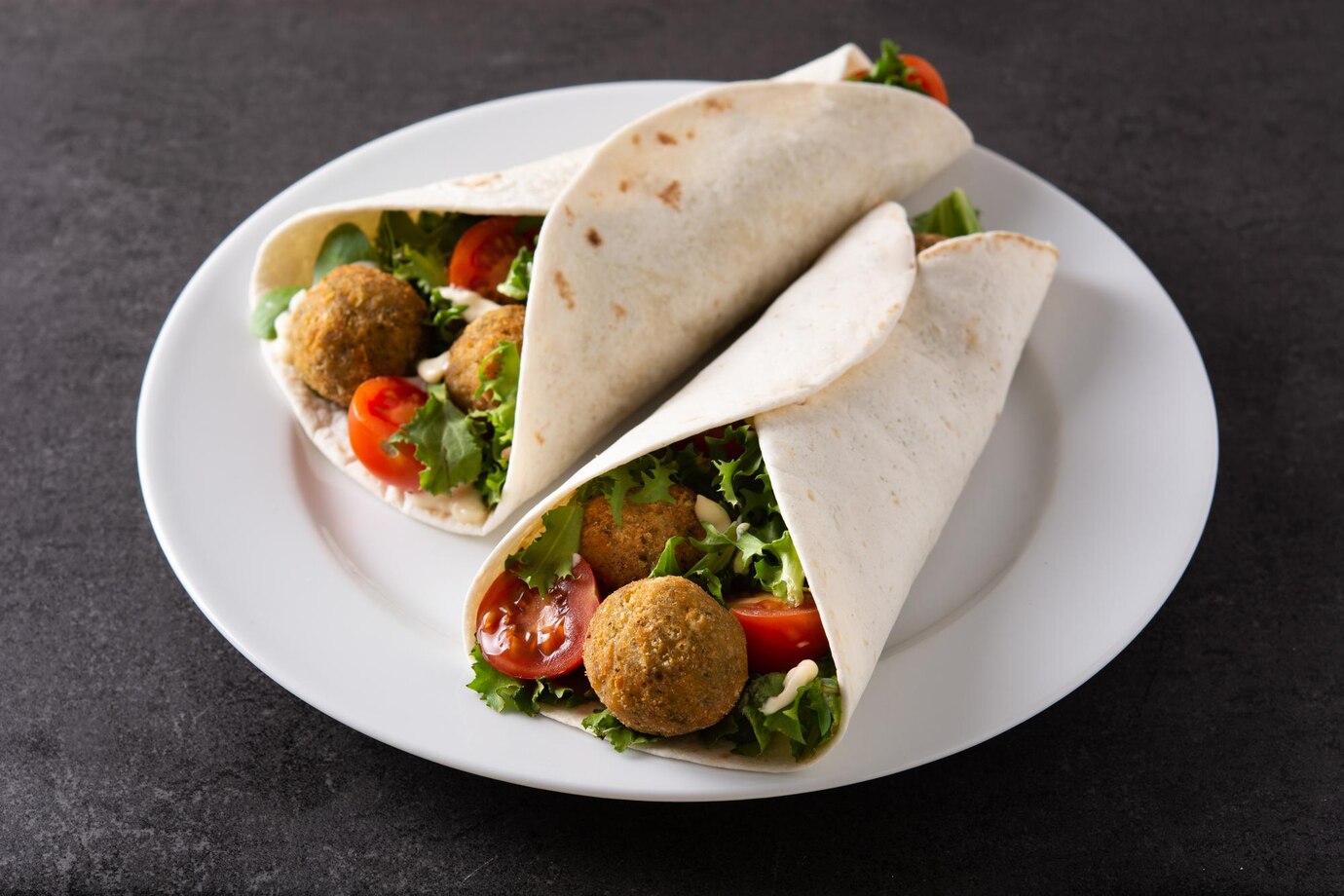 Two falafel wraps with tomatoes and lettuce on a white plate against a dark background.