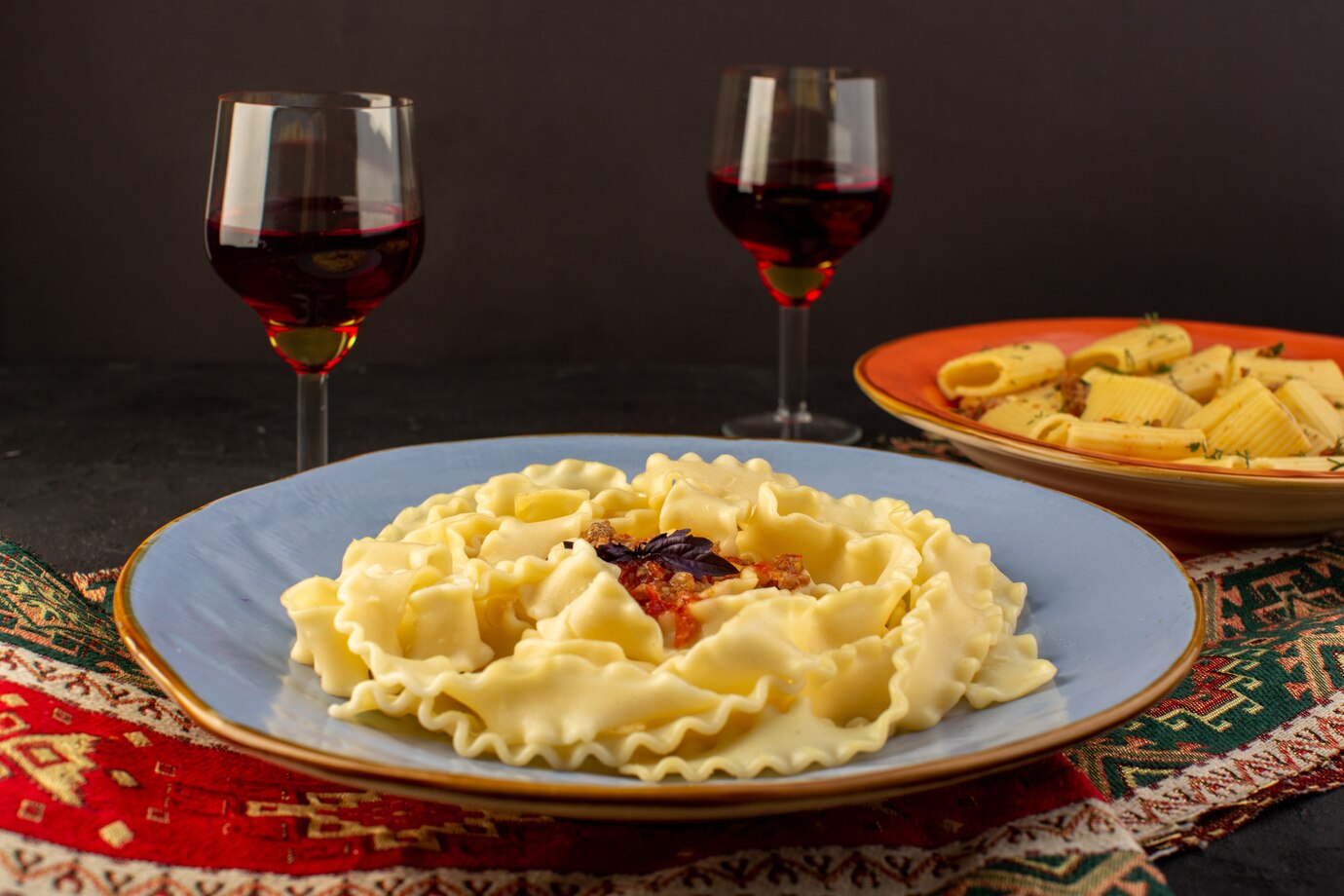Plate of ravioli with tomato sauce, garnished with basil, accompanied by two glasses of red wine and another pasta dish in the background.